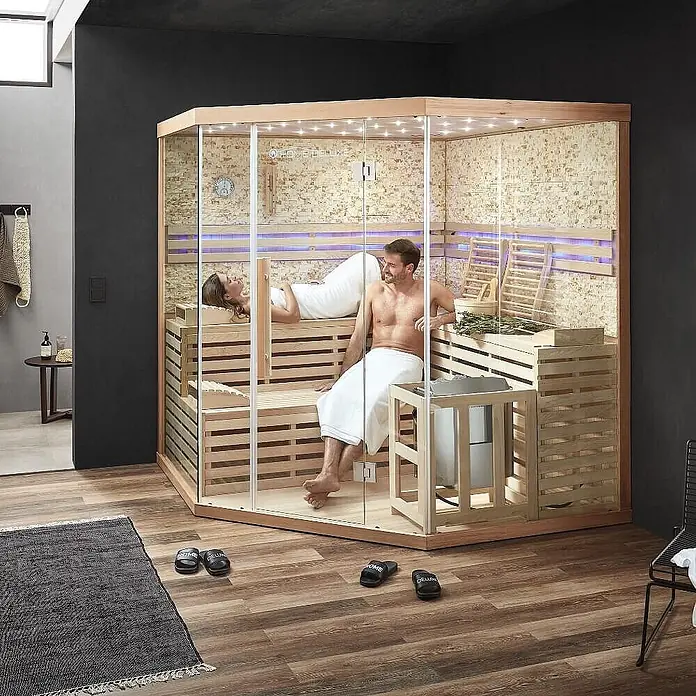 Home Deluxe Elementsauna SKYLINE BIG XL KUNSTSTEINWAND - Elektroofen, Steuerung Ofen: Integriert, L x B x H: 200 x 200 x 210 cm - Moderne Indoor-Saunakabine aus hellem Holz und Glas, mit zwei Personen beim Entspannen.