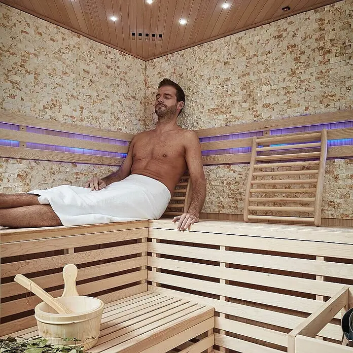 Home Deluxe Elementsauna SKYLINE BIG XL KUNSTSTEINWAND - Elektroofen, Steuerung Ofen: Integriert, L x B x H: 200 x 200 x 210 cm - Mann entspannt in moderner Sauna mit Holzliegen, Steinwänden und blauen LEDs. Saunaeimer und Kelle sichtbar.