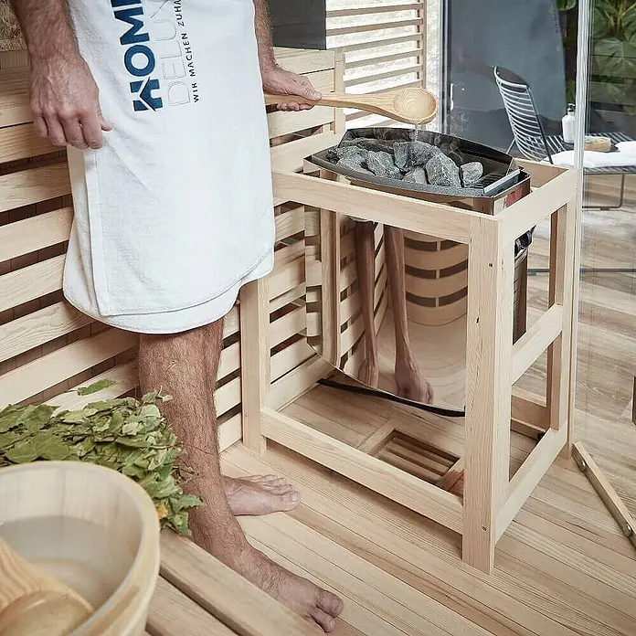 Home Deluxe Elementsauna SKYLINE BIG XL KUNSTSTEINWAND - Elektroofen, Steuerung Ofen: Integriert, L x B x H: 200 x 200 x 210 cm - Holzsaunaofen mit Metallschale und grauen Steinen, in Gebrauch, wobei Wasser aus einer Kelle gegossen wird.