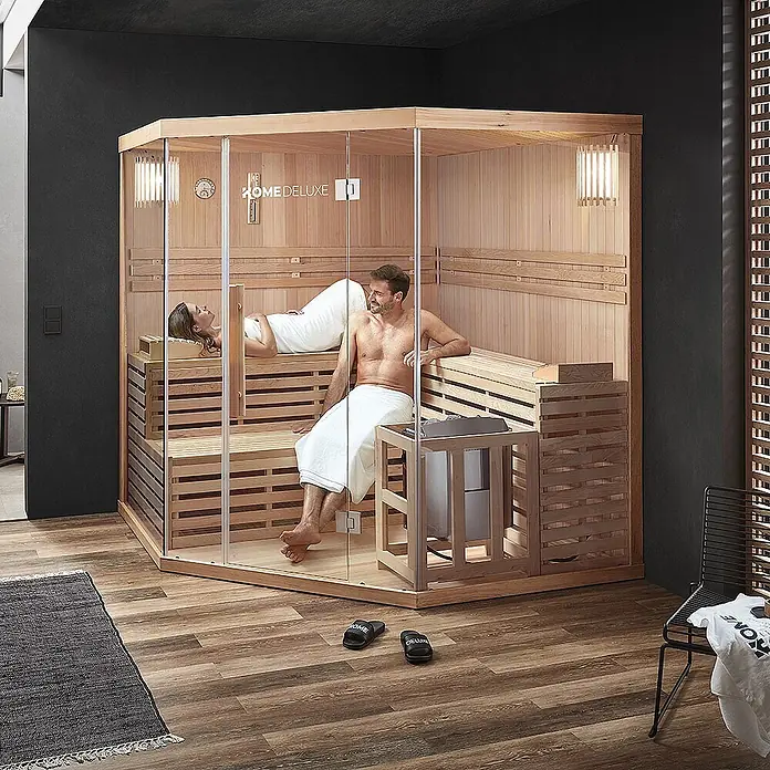 Home Deluxe Elementsauna SKYLINE BIG XL - Elektroofen, Steuerung Ofen: Integriert, L x B x H: 200 x 200 x 210 cm - Ecksauna aus hellem Holz mit Glaswänden, von einem Paar genutzt.