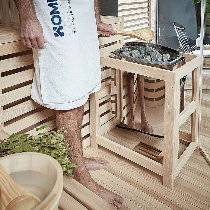 Home Deluxe Elementsauna SKYLINE BIG XL - Elektroofen, Steuerung Ofen: Integriert, L x B x H: 200 x 200 x 210 cm - Saunaofen mit heißen Steinen in hellem Holzrahmen, Person gießt Wasser für Dampf auf.