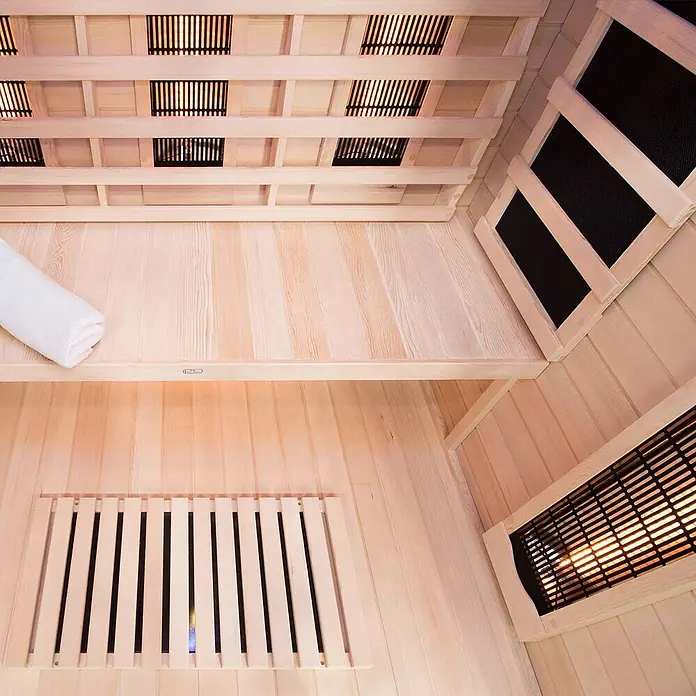 Home Deluxe Infrarotkabine SAHARA L -  - Heller Holzkabinen-Innenraum einer Infrarotsauna mit leuchtenden Heizstrahlern, Sitzbank und gefaltetem weißem Handtuch.