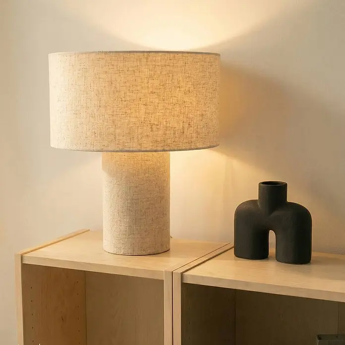 Lindby Tischleuchte Elfie - L x B x H: 35 x 35 x 40 cm, creme / amber, E27 - Eine hohe Tischlampe mit hellem Stoffschirm und zylindrischem Fuß, die warmes Licht auf einem Holzregal spendet.