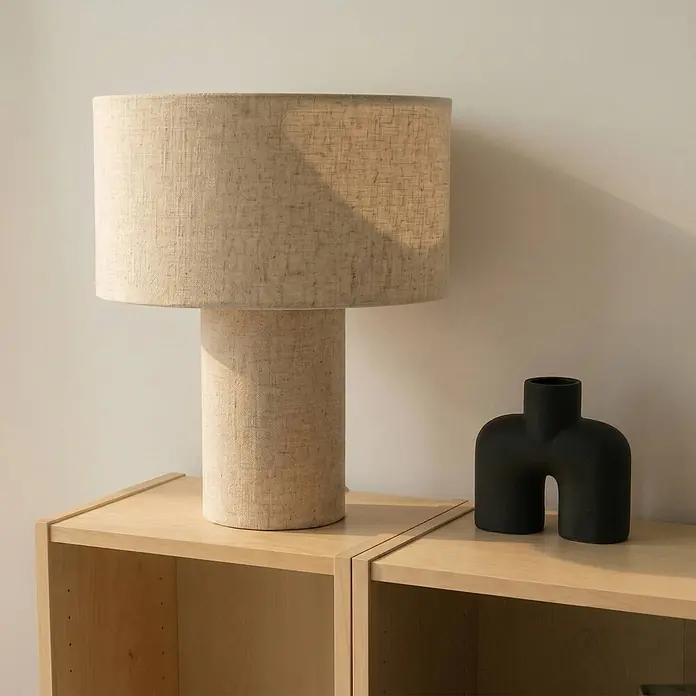 Lindby Tischleuchte Elfie - L x B x H: 35 x 35 x 40 cm, creme / amber, E27 - Zylindrische Tischleuchte aus Naturleinen auf hellen Holzregalen, daneben eine matte, schwarze abstrakte Vase.