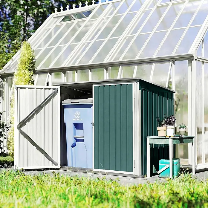Outsunny Mülltonnenbox - Stahl - Dunkelgrüner Metall-Geräteschuppen mit offener weißer Tür, dahinter eine blaue Wertstofftonne, neben einem Gewächshaus und einem kleinen Tisch.