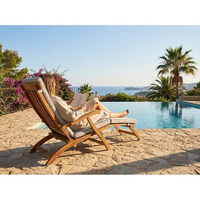 Prestige Home Living Outdoor Sonnenliege - B x T x H: 60 x 190 x 95 cm, Akazie, Braun - Holzliege mit hellen Polstern, von einer Frau zum Entspannen und Lesen am Pool mit Meerblick genutzt.