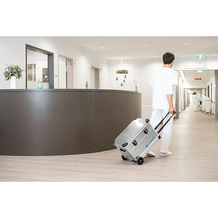 Zarges Aufbewahrungsbox K 424 XC Mobil Universal Alu Box 105l -  - Silberner ZARGES Aluminiumkoffer auf Rollen, der von medizinischem Personal durch einen Gang gezogen wird.
