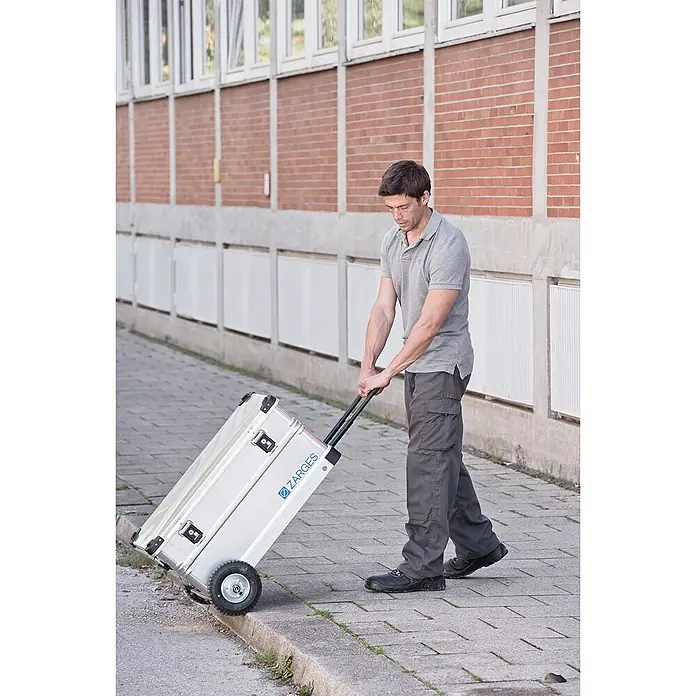 Zarges Aufbewahrungsbox K 424 XC Mobil Universal Alu Box 105l -  - Mann zieht einen silbernen Zarges Aluminiumkoffer mit Rädern auf einem Gehweg, der seine Tragbarkeit zeigt.