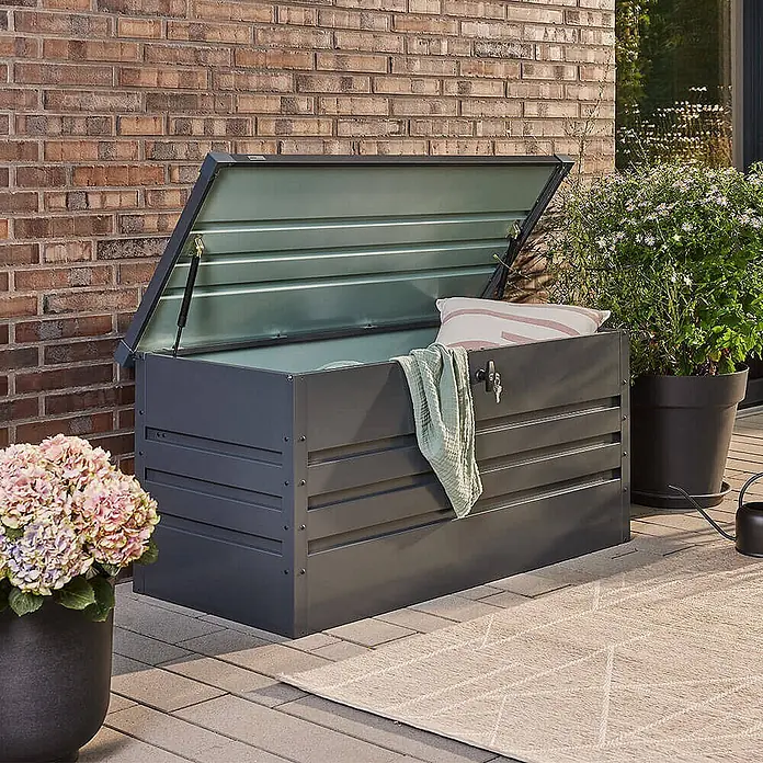 Home Deluxe Garten-Aufbewahrungsbox MEGABOX XXL Größe: 600L - L x B x H: 158 x 64 x 62 cm, Anthrazitgrau, Stahl - Dunkelgraue Outdoor-Aufbewahrungsbox mit geöffnetem Deckel, der ein Kissen und eine Decke zeigt. Steht auf einer Terrasse neben Pflanzen.