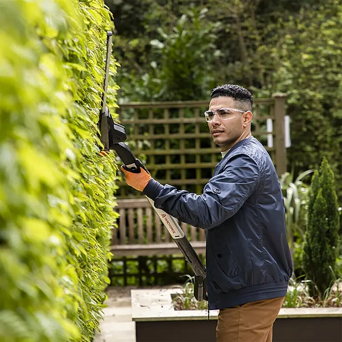 Yard Force Akku-Heckenschere iFlex LH F35 - 1 Akku, Schnittlänge: 35 cm - Mann mit Schutzbrille und Handschuhen trimmt mit schwarzem und weißem Heckenschneider eine grüne Hecke.