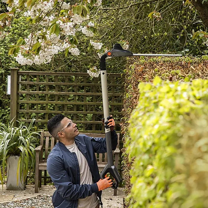 Yard Force Akku-Heckenschere iFlex LH F35 - 1 Akku, Schnittlänge: 35 cm - Mann schneidet eine hohe Hecke mit einer grauen und schwarzen Langstiel-Heckenschere.