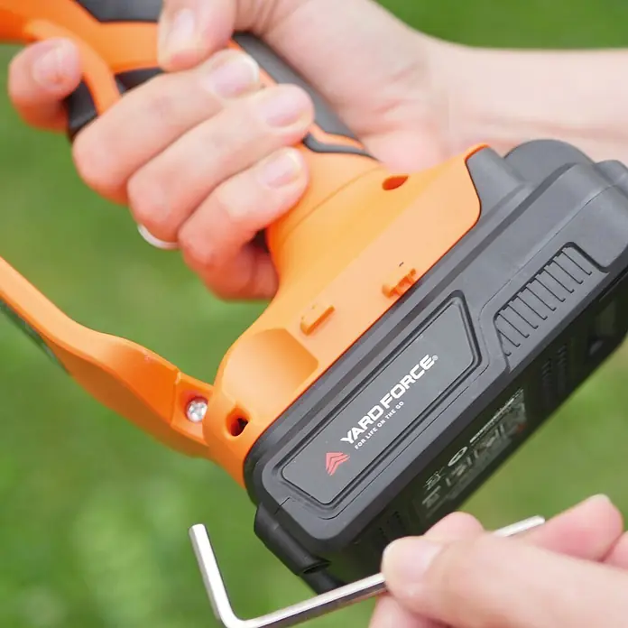 Yard Force Einhand-Akku-Kettensäge LS C13 - 2 Ah, 1 Akku - Person hält orange-schwarzes YARD FORCE Elektrowerkzeug; ein Inbusschlüssel wird zur Einstellung an der Basis gehalten.