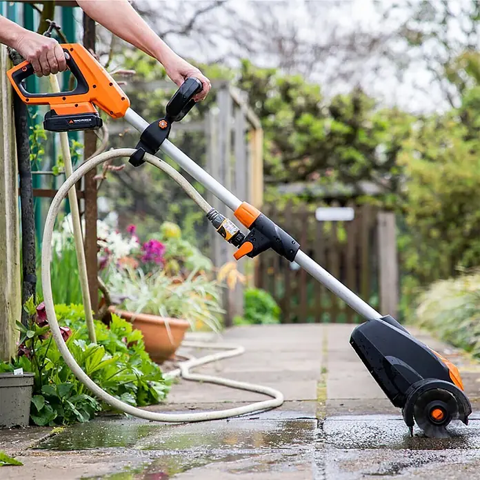 Yard Force Akku-Reinigungsbürste LW CPC1 - 20 V, 1 Akku, 4 Ah - Person reinigt eine nasse Pflasterterrasse mit einem orange-schwarzen Akku-Terrassenreiniger und Schlauch.
