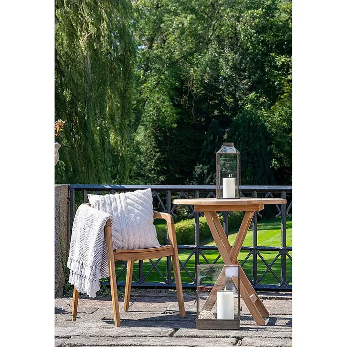 Ebuy24 Gartenstuhl Otero 2er-Set - 2 Stk., Massiv Teakholz, Natur, B x T x H: 52 x 55 x 76 cm - Sitzgruppe im Freien mit Holzstuhl, weißem Kissen und Decke neben einem kleinen Holztisch. Zwei Laternen mit Kerzen schaffen eine gemütliche Terrassenatmosphäre.