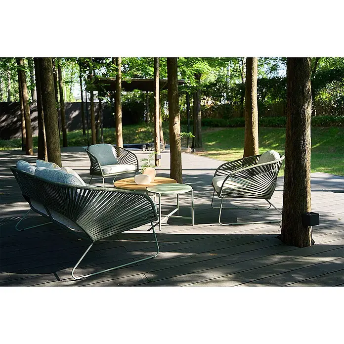 Ebuy24 Loungemöbel-Set Novella - 5 -tlg., Schaum, Polyester, Stahl, Grau - Dunkelgrünes geflochtenes Outdoor-Sofa und zwei Sessel mit hellen Polstern, begleitet von zwei Tischen auf einer Holzterrasse zwischen Bäumen.