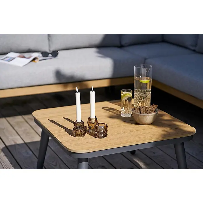 Ebuy24 Loungemöbel-Set Camilla - 4 -tlg., Nonwood (PE), Aluminium, Polyester, Graubeige - Zwei bernsteinfarbene Glaskerzenhalter mit brennenden weißen Kerzen auf einem Holztisch im Freien.