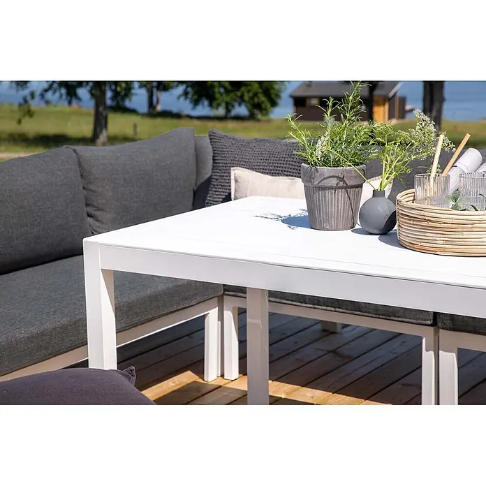 Ebuy24 Loungemöbel-Set Ramos - 3 -tlg., Aluminium, Grau - Weißer Outdoor-Esstisch mit grauen Polstern, auf einer Holzterrasse mit Pflanzen und Getränken.