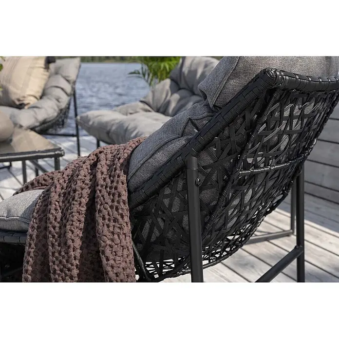 Ebuy24 Loungemöbel-Set Tina - 4 -tlg., Stahl, Schwarz - Schwarzer Outdoor-Loungesessel mit grauen Polstern und brauner Waffelstrickdecke auf einer Holzterrasse am Wasser.
