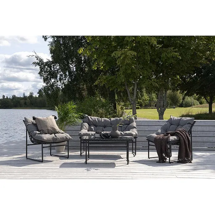 Ebuy24 Loungemöbel-Set Tina - 4 -tlg., Stahl, Schwarz - Outdoor-Lounge-Set mit dunklen Gestellen und plüschigen grauen Kissen, bestehend aus Sofa, zwei Sesseln und Couchtisch, auf einer Terrasse am See.
