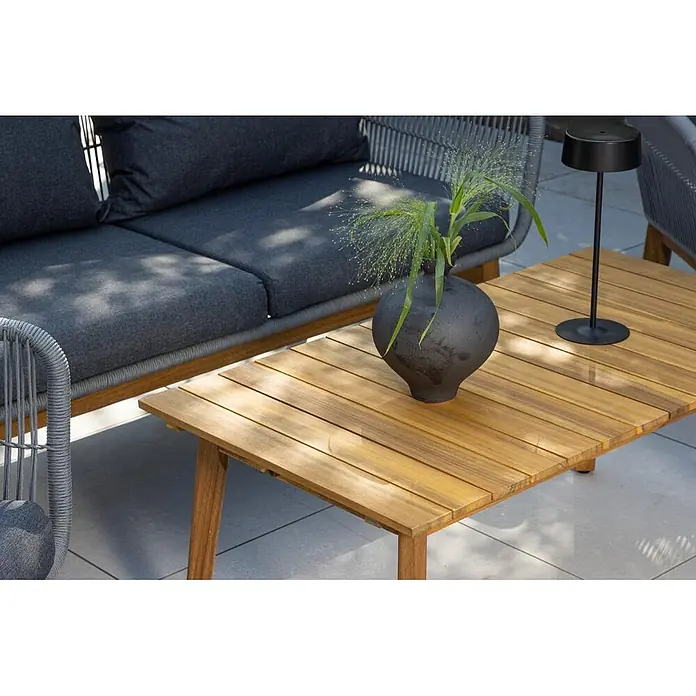 Ebuy24 Loungemöbel-Set Chania - 4 -tlg., Akazie, Grau - Heller Lattenholz-Couchtisch auf einer Terrasse, mit dunkler Vase mit Grün und schwarzer Lampe, neben einem grauen Outdoor-Sofa.