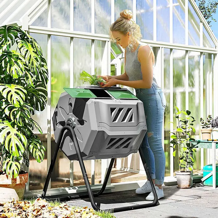 Outsunny Komposter - 160 l, 71 x 65 x 96 cm - Eine Frau füllt organische Abfälle in einen grau-grünen, drehbaren Komposter mit zwei Kammern im Garten vor einem Gewächshaus.