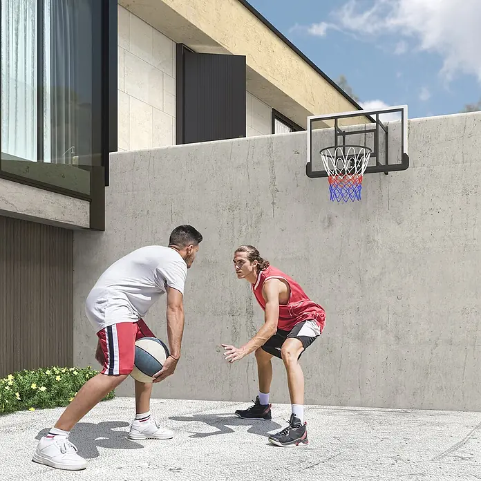 Sportnow Basketballkorb - Stahl, Schwarz - Schwarzer Basketballkorb mit klarem Backboard, weißem Rahmen und rot-blau-weißem Netz, an Betonwand, wo zwei Männer spielen.