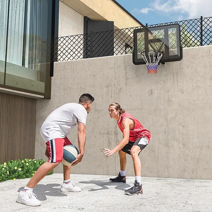 Sportnow Basketballkorb - Stahl, Schwarz - Schwarzer Basketballkorb mit transparenter Platte und buntem Netz, an einer Betonwand befestigt, zwei Männer spielen draußen.