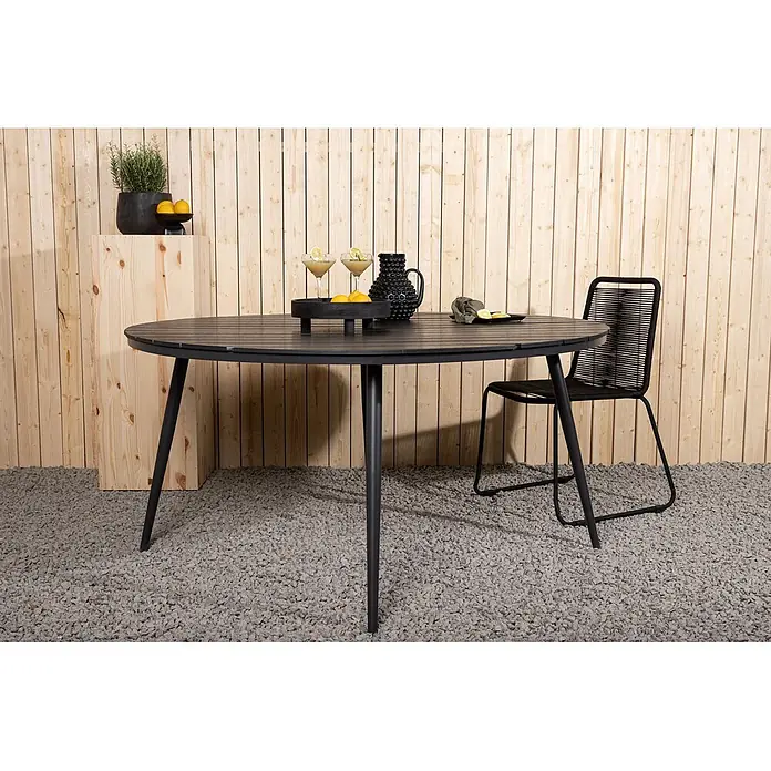 Ebuy24 Gartentisch rund Break - Ø x H: 150 x 74 cm, Aluminium, Schwarz, Stapelbar - Ein schwarzer, ovaler Lamellen-Esstisch für den Außenbereich mit schwarzen Beinen und passendem Stuhl auf Kies. Mit Getränken und Zitronen dekoriert.