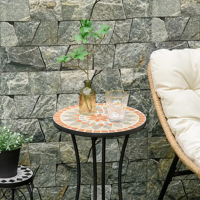 Outsunny Garten-Beistelltisch rund - Ø x H: 35,5 x 53,5 cm, Keramik, Stahl, Schwarz - Runder Mosaik-Bistrotisch mit orangefarbenen, beigen und grauen Fliesen, mit Pflanze und Glas, draußen neben einem Stuhl.