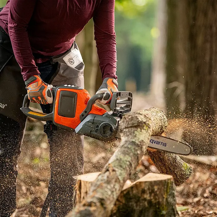 Husqvarna Akku-Kettensäge - 36 V, 4 Ah, Li-Ionen, 1 Akku, Schwertlänge: 35 cm - Eine Person in Schutzkleidung sägt mit einer orange-grauen Husqvarna Kettensäge einen Baumstamm, Sägespäne fliegen.