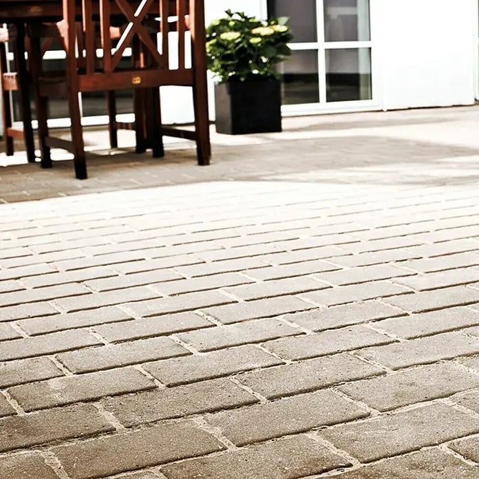 Dansand Fugensand No GrowSand/Neutral, 20 kg - Detailaufnahme eines gepflasterten Außenbereichs mit braunen Pflastersteinen, Sitzmöbeln und Pflanzen. Harmonische Gestaltung für den Garten.