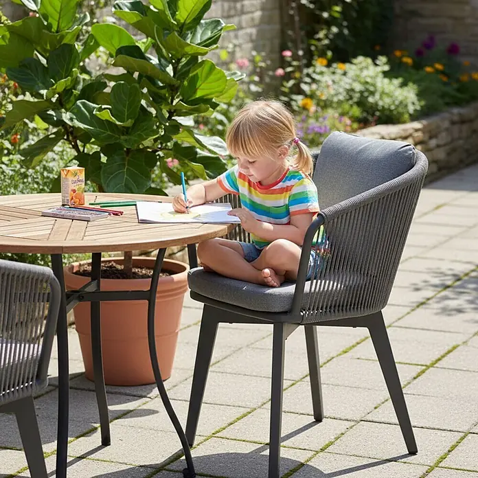 Bene Living Gartenstuhl-Set Marbella - 2 Stk., Aluminium mir Rope-Geflecht, grau, B x T x H: 63 x 58 x 78 cm - Kind zeichnet an einem runden Holztisch im Freien mit einem grauen, geflochtenen Esszimmerstuhl und Kissen.