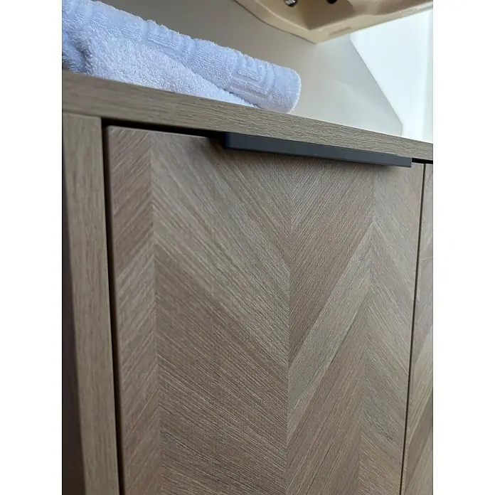 Schildmeyer Waschbeckenunterschrank Piazza - B x T x H: 651 x 330 x 536 mm, Eiche Hell/Eiche Chevron - Heller Holzschrank mit Fischgrätmuster, dunklem Griff und weißem Handtuch.