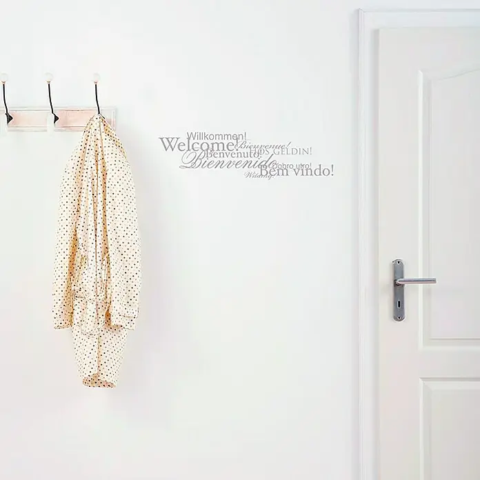 Komar Dekosticker Welcome - Silber, 50 x 70 cm - Wandtattoo mit Willkommen in verschiedenen Sprachen als Dekoration, neben einer Tür und einer Garderobe.