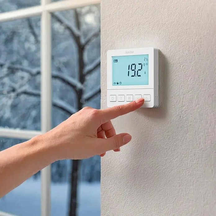 Hama Raumthermostat 00176629 Unterputz per App/Sprache steuern - Weiß - Weißes Hama Smart-Thermostat an der Wand, zeigt 19.2°C, eine Hand bedient es vor einem verschneiten Fenster.