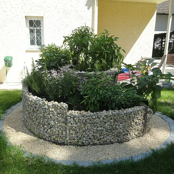 Min2C Bruchsteine - Gelb, Körnung: 30 mm - 60 mm, 25 kg - Gabionen-Kräuterspirale mit verschiedenen Kräutern und Pflanzen befüllt, umrandet von Kies und Rasen im Garten.