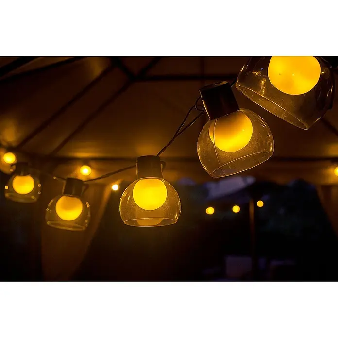 Garza Guirnalda luminosa solar Helios - Para exterior, Blanco cálido, 7 m, Número de bombillas: 10 ud., Con energía solar - Guirnalda de luces amarilla cálida con bombillas de globo de plástico transparente colgando bajo un toldo oscuro, iluminando un espacio exterior de noche.