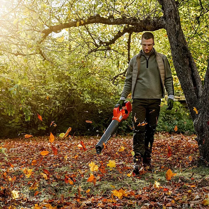 Husqvarna Akku-Laubbläser 230iB - 1 Akku, Blasgeschwindigkeit: 220 km/h - Mann im Garten nutzt einen rot-schwarzen Laubbläser, um Herbstlaub zu entfernen.