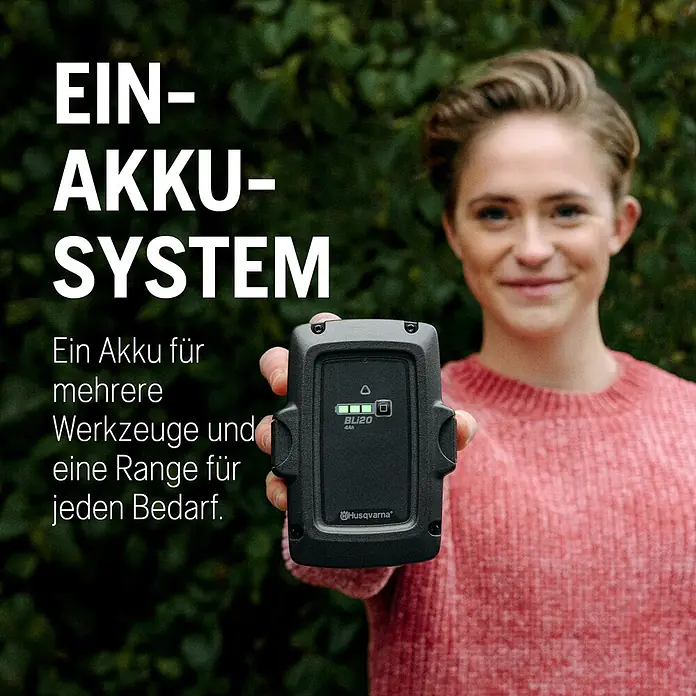 Husqvarna Akku-Laubsauger & -Laubbläser 120iBV - Ohne Akku, Blasgeschwindigkeit: 172 km/h - Lächelnde Person hält einen schwarzen Husqvarna BLi20 4Ah Akku, betont dessen „Ein-Akku-System“ für verschiedene Werkzeuge.