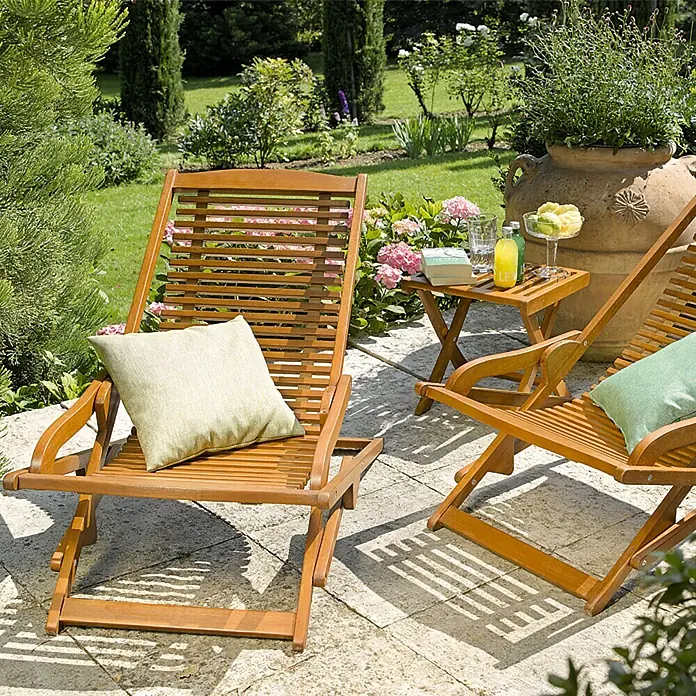 Sunfun Diana Tumbona reclinable - Marrón, Eucalipto - Dos tumbonas de jardín de madera con cojines y una pequeña mesa con bebidas en un jardín pintoresco.