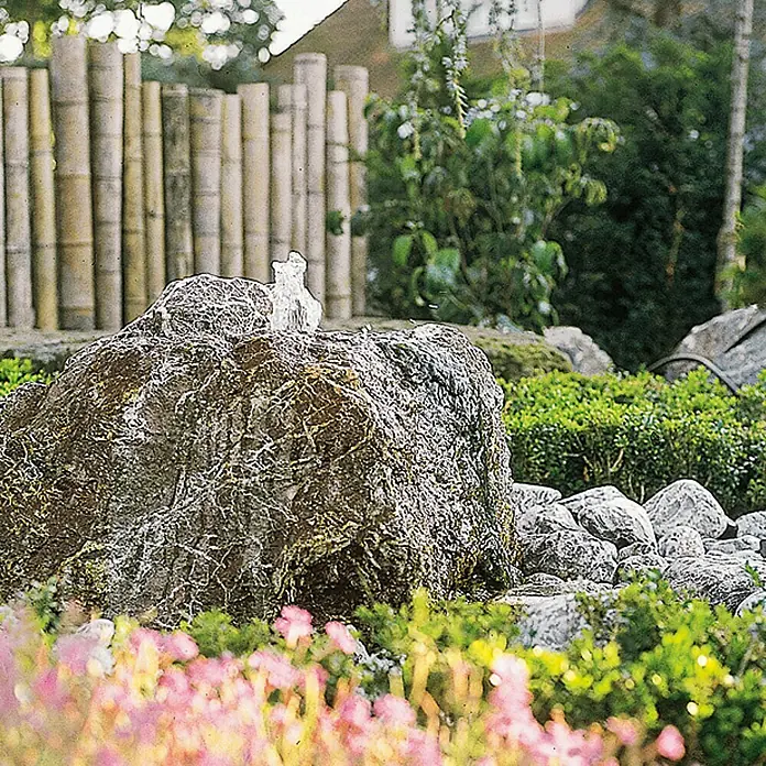 Oase Statuen-Zimmerbrunnenpumpe Aquarius Universal 440 I - 5 W, Max. Fontänenhöhe: 75 cm - Nahaufnahme eines steinernen Gartenbrunnens mit Wasserfontäne, umgeben von Blumen und Bambuszäunen für eine ruhige Gartengestaltung.