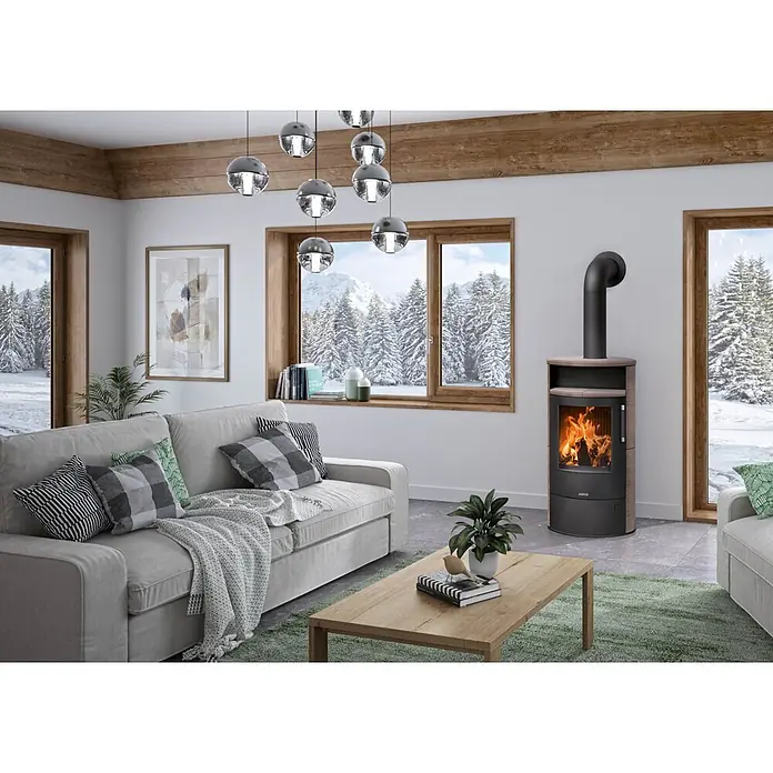 Justus Kaminofen Island 7 2.0 - 7 kW, Raumheizvermögen: 144 m³, Farbe Verkleidung: Grappa - Ein moderner dunkelgrauer Kaminofen mit heller Holzplatte und Seitenpaneelen, aktiv brennend, in einem gemütlichen Wohnzimmer mit Bergblick.