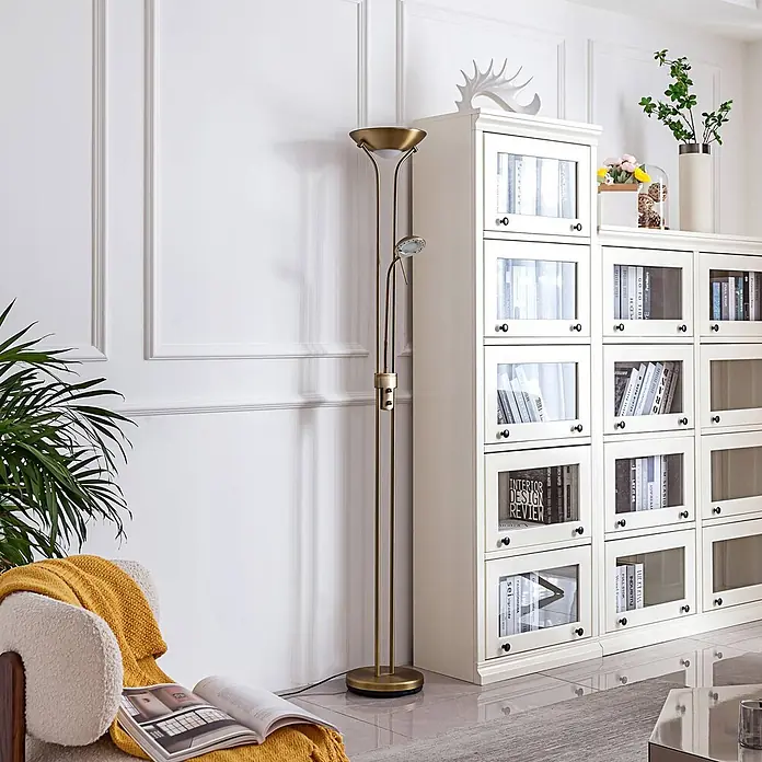 Lindby LED-Deckenfluter Josefin - Höhe: 180 cm, bronze / altmessing, bronze / altmessing, Warmweiß - Eine messingfarbene Stehlampe mit Deckenfluter und Leselicht steht neben einem weißen Bücherregal und Sessel.