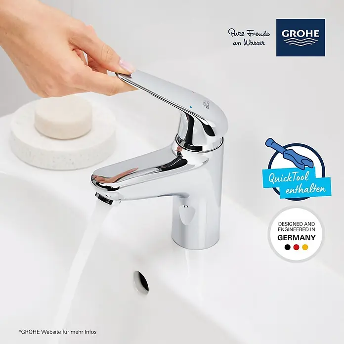 Grohe Swift Waschtischarmatur M-Size - Ausladung Armatur: 116 mm, Chrom, Glänzend - Ein verchromter Badarmatur in einem weißen Waschbecken, eine Hand dreht das Wasser auf. Eine Seife liegt daneben.