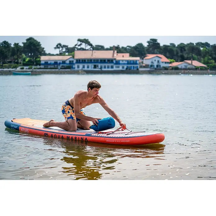 Aqua Marina SUP-Board-Set Atlas - L x B x H: 366 x 86 x 15 cm, Tragkraft: 180 kg, Orange - Ein orange-blaues aufblasbares Aqua Marina Stand-Up-Paddleboard mit einem Mann, der im Wasser kniet und eine blaue Trockentasche verstaut.