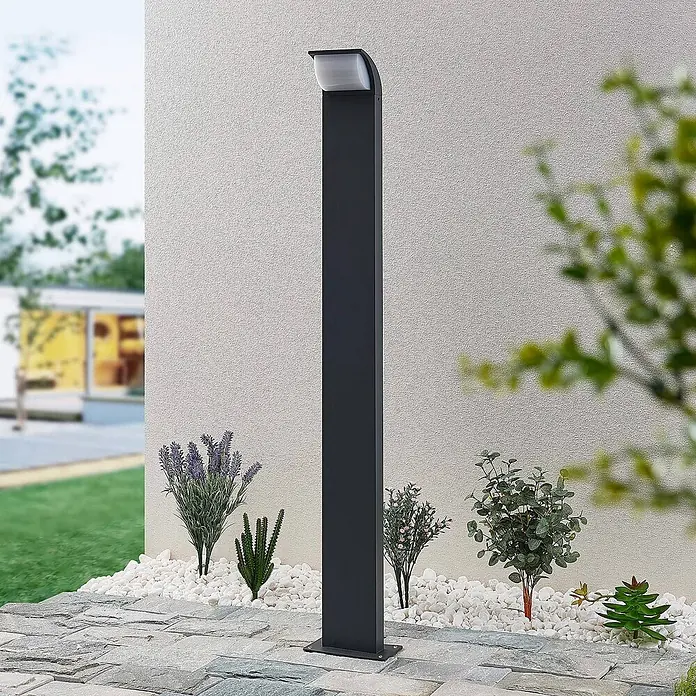 Lucande Außenlaterne Tinna -  - Dunkelgraue Außen-Pollerleuchte mit weißem Lichtelement, auf einer Terrasse zwischen Pflanzen und weißem Kies installiert.