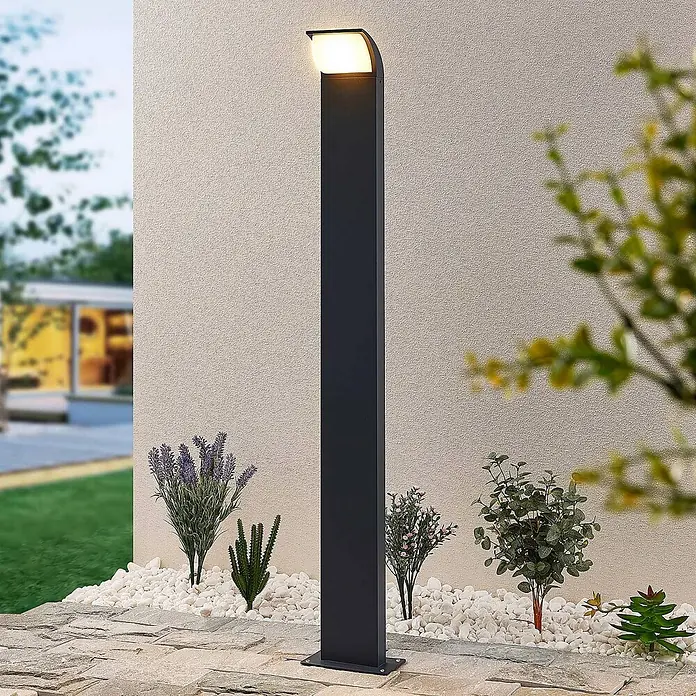 Lucande Außenlaterne Tinna -  - Dunkelgraue moderne Wegeleuchte mit beleuchteter weißer Oberseite, stehend in einem Gartenbeet mit weißen Kieselsteinen und Pflanzen.