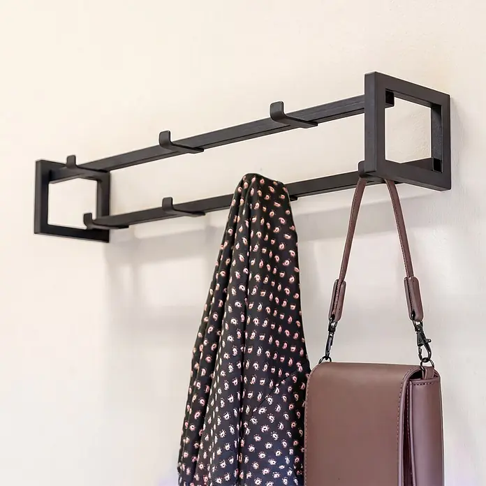 Spinder Design Garderobenleiste Axel 1 - L x B x H: 11,5 x 60 x 10 cm, Anzahl Haken: 7 Stk., Schwarz - Schwarzmetallene Garderobe mit schwarzem gemustertem Kleidungsstück und brauner Handtasche.