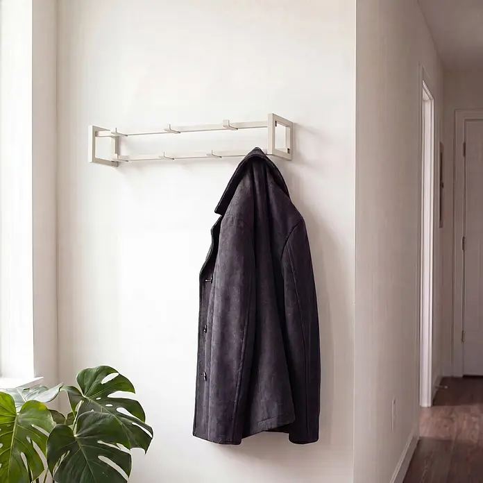 Spinder Design Garderobenleiste Axel 1 - L x B x H: 11,5 x 60 x 10 cm, Anzahl Haken: 7 Stk., Weiß - Ein minimalistischer, heller Garderobenständer mit Haken, der einen dunkelgrauen Mantel in einem Flur hält.