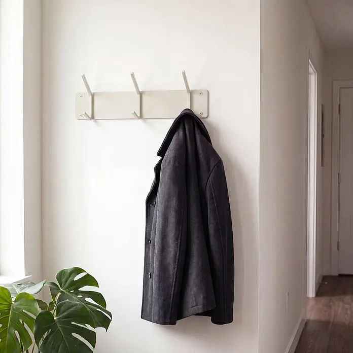 Spinder Design Garderobenleiste Cappa - L x B x H: 1,6 x 40 x 12 cm, Anzahl Haken: 3 Stk., Weiß - Dunkelgrauer Mantel hängt an einer hellbeigen Wandgarderobe mit vier Haken in einem hellen Raum mit Pflanze.